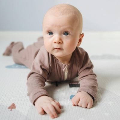 Bebé deitado de barriga para baixo com fato castanho claro sobre tapete branco com padrões coloridos