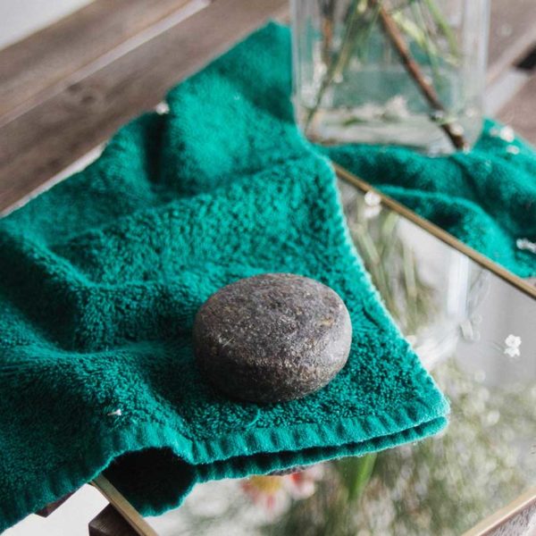 CHAMPÔ SÓLIDO MIND THE TRASH – CABELOS OLEOSOS Saco verde com pedra redonda cinzenta sobre superfície de madeira com vidro e plantas ao fundo