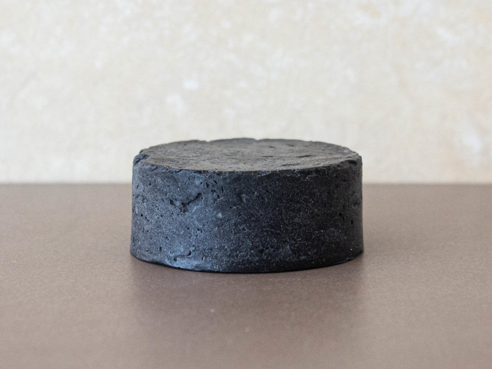 Champô sólido para Homem All-In-One - Miristica Barra sólida preta com textura porosa sobre superfície castanha lisa.
