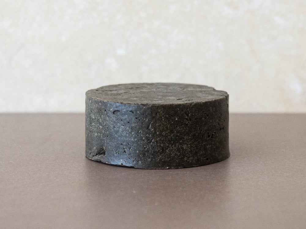 Champô sólido para cabelos oleosos e caspa - Miristica Produto cilíndrico preto com textura rugosa sobre superfície castanha