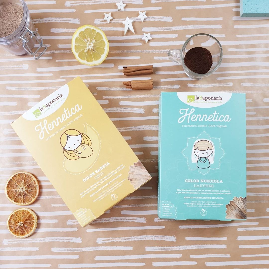 Tintas de Cabelo 100% Vegetal – várias cores disponíveis Duas caixas de coloração natural do cabelo la sponaria Hennetica, uma amarela e uma azul, com ingredientes naturais ao redor sobre superfície listrada.