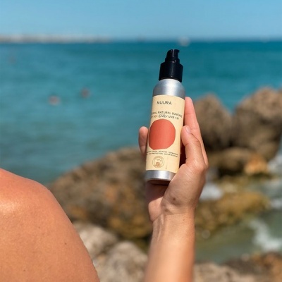 Frasco de protetor solar NUURA segurado por mão à beira-mar com pedras ao fundo.