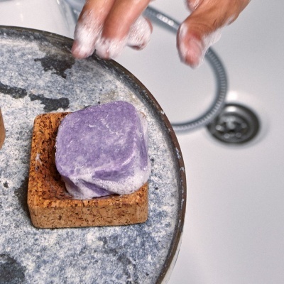 Barra de sabão roxa em saboneteira de cortiça sobre mesa cinza com mãos e ralo ao fundo