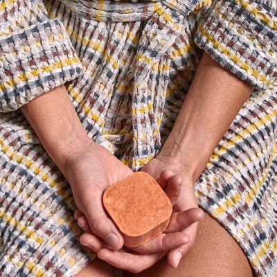 Mãos femininas a segurar sabonete sólido laranja, vestindo roupão almofadado listrado tons claros