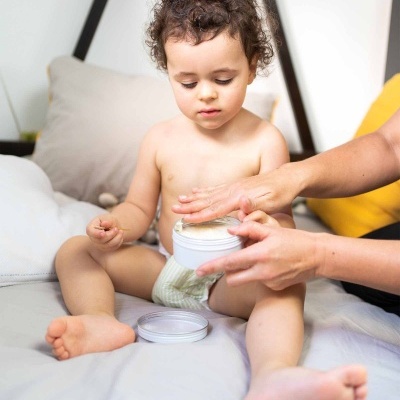 Bebé com fralda e adulto a aplicar creme em recipiente branco