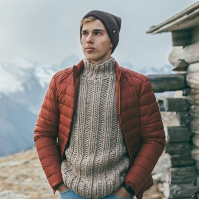 Homem com casaco acolchoado vermelho, suéter bege de malha e gorro cinza junto a cabana de madeira e montanhas