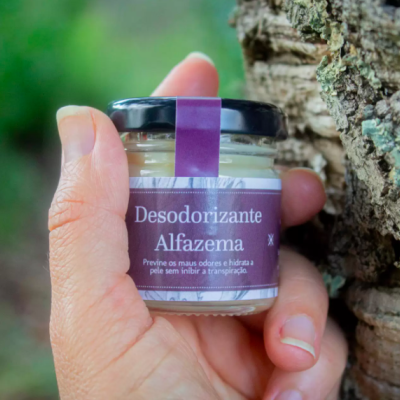 Frasco pequeno de vidro com desodorizante de alfazema e rótulo roxo segurado junto a um tronco de árvore