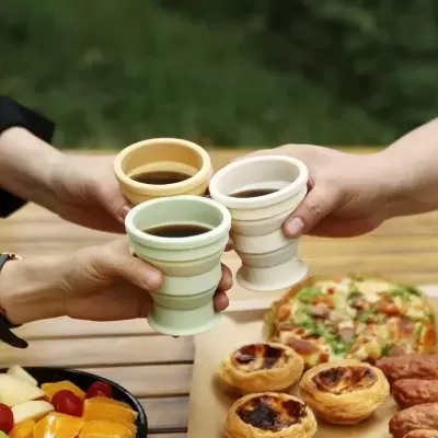 Três copos de cerâmica coloridos com bebida escura brindando sobre mesa com pizza e pastéis de nata