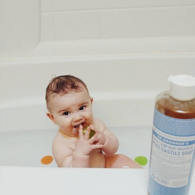 Bebé numa banheira a morder brinquedo verde com garrafa de sabão Dr. Bronner's ao lado