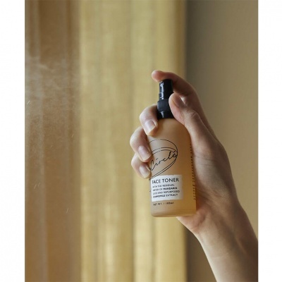 Frasco de spray de toner facial translúcido amarelo com tampa preta na mão em frente a cortina bege