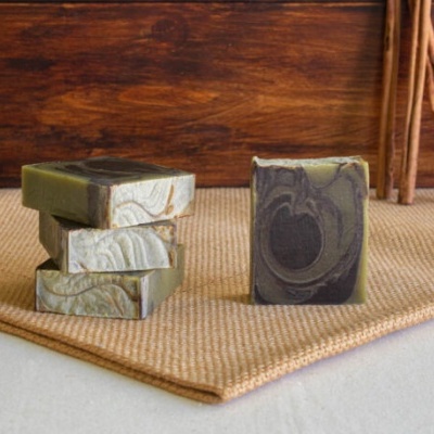 Barras de sabonete marmoreadas em verde, bege e preto sobre tapete bege, com fundo de madeira e pauzinhos de canela