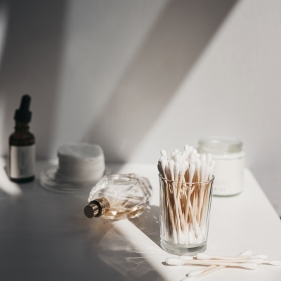 Acessórios de higiene e beleza numa superfície branca com cotonetes, perfume, discos de algodão e frascos.