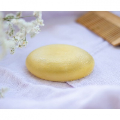 Sabonete amarelo redondo sobre tecido lilás com flores brancas e pente de madeira ao fundo