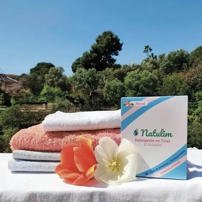 Detergente Natulim em caixa branca e azul com toalhas coloridas e flores sobre mesa com fundo de árvores
