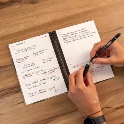 Agenda semanal aberta com anotações e caneta preta Infinitebook na mão de uma pessoa sobre mesa de madeira clara