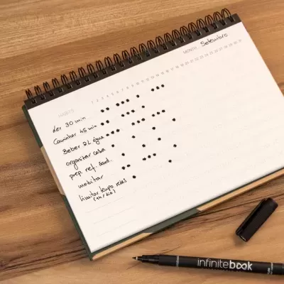 Caderno de hábitos semanal aberto com caneta preta da marca infinitebook sobre mesa de madeira clara