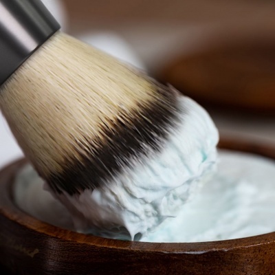 Pincel de barbear a aplicar creme azul numa taça de madeira