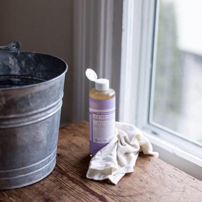 Frasco de sabão líquido com rótulo roxo e pano branco ao lado de balde metálico junto a janela