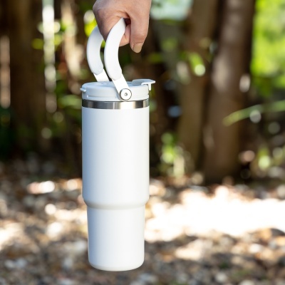 Garrafa térmica branca com tampa e alça segurada numa mão, exterior com vegetação