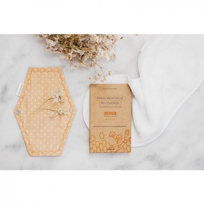Pensa menstrual reutilizável laranja com bolinhas, embalagem em papel kraft e pano branco sobre mesa clara com flores secas.