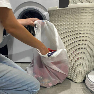 Pessoa a colocar roupa branca numa bolsa de pano transparente junto a cesta de roupa e máquina de lavar