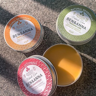 Três latas redondas de desodorizante natural nas cores laranja, verde e rosa sobre superfície cinza