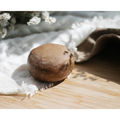 Barra redonda de sabão marrom sobre pano branco e madeira clara com flores ao fundo