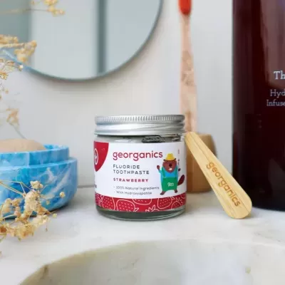 Pasta de dentes sólida georganics sabor morango em frasco de vidro com tampa metálica e espátula de madeira sobre bancada de mármore