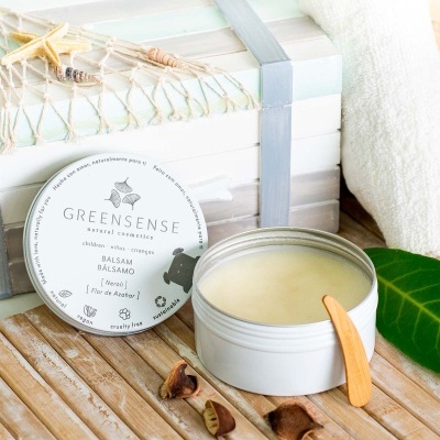 Bálsamo em pote branco aberto com espátula de madeira junto a caixa e toalha branca sobre superfície de madeira.