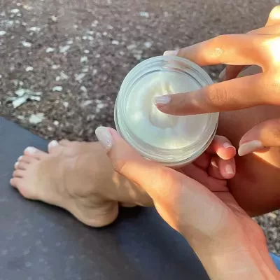 Pote transparente com creme branco e mão tocando o creme, fundo com chão de gravilha e pé descalço