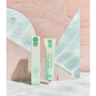 Tubo e caixa do protetor solar ODYSKIN SPF 50 em superfície clara com fundo rosa texturizado e triângulos azulados.