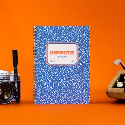 Caderno azul com padrão branco em espiral e etiqueta 'INFINITE BOOK' sobre fundo laranja com máquina de escrever, caneta e câmara fotográfica ao lado