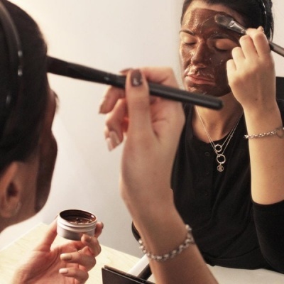 Mulher aplica máscara facial castanho escuro no rosto com pincel frente a espelho