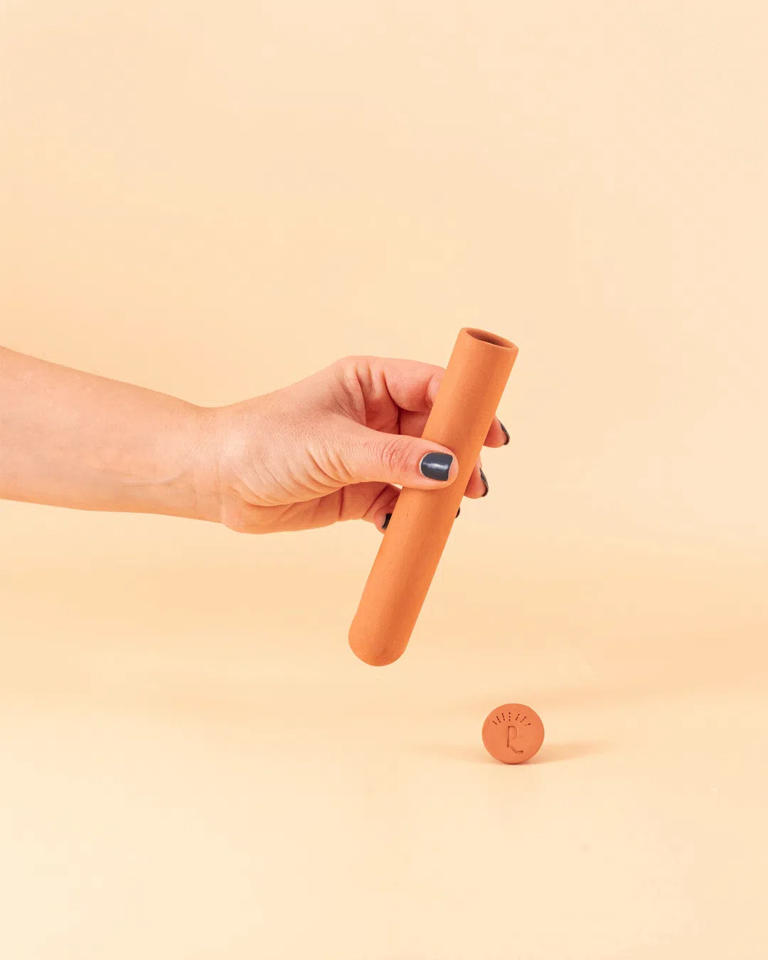 Tubo Barro para Rega Estojo cilíndrico terracota com tampa gravada, segurado por mão no fundo bege