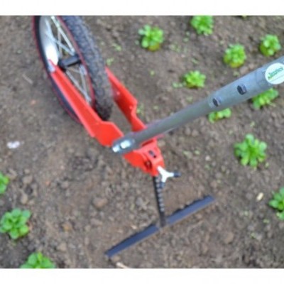 Enxada de mão com cabo metálico cinzento e cabeça preta ao lado de roda vermelha em solo de terra com plantas verdes