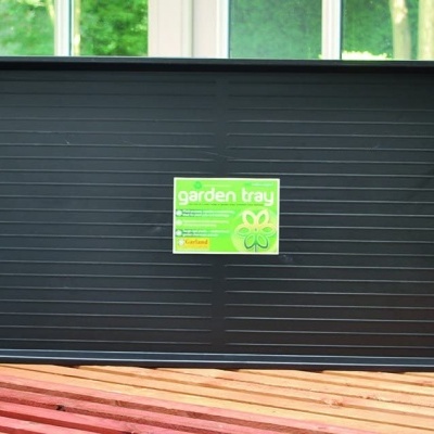 Tabuleiro de cultivo preto com etiqueta verde e texto garden tray sobre mesa de madeira com plantas ao fundo