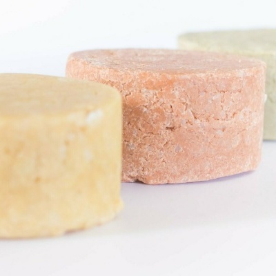 Três barras redondas de shampoo sólido em cores bege, rosa claro e verde claro sobre fundo branco