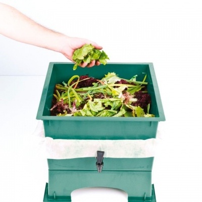 Compostor de cozinha verde com resíduos orgânicos e mão a depositar folhas