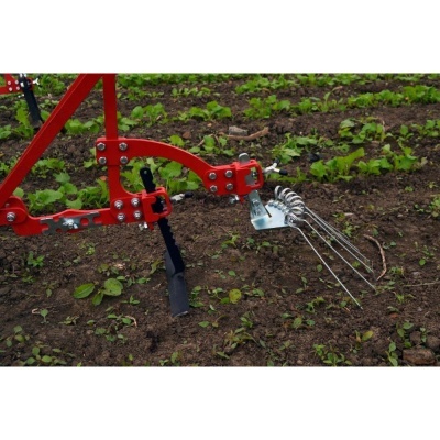 Ferramenta agrícola vermelha com garras metálicas em solo arado e plantas pequenas