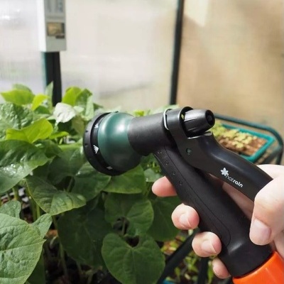 Regador manual preto e verde com cabo laranja a regar plantas verdes numa estufa