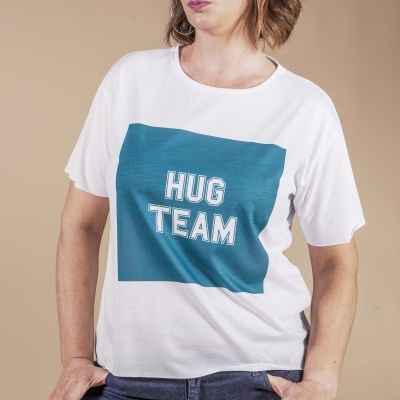 T-shirt branca com estampa azul e texto HUG TEAM, usada com calças de ganga num fundo bege.