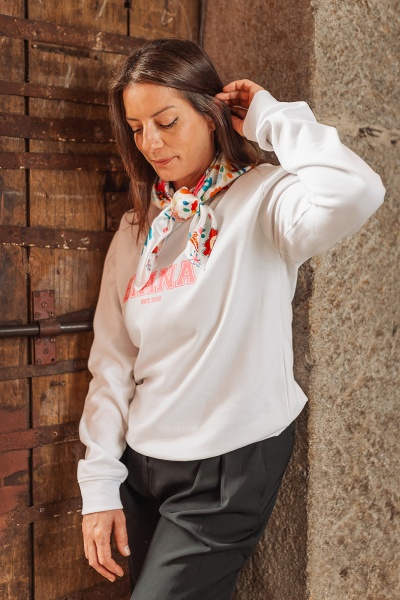 Mulher com sweatshirt branca e lenço colorido junto a parede de pedra e porta de madeira.