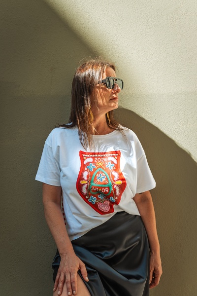 Mulher a usar t-shirt branca com estampa colorida e saia preta encostada numa parede bege