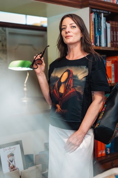 Mulher de camisa preta com estampa frontal e saia branca em biblioteca com estante de livros ao fundo