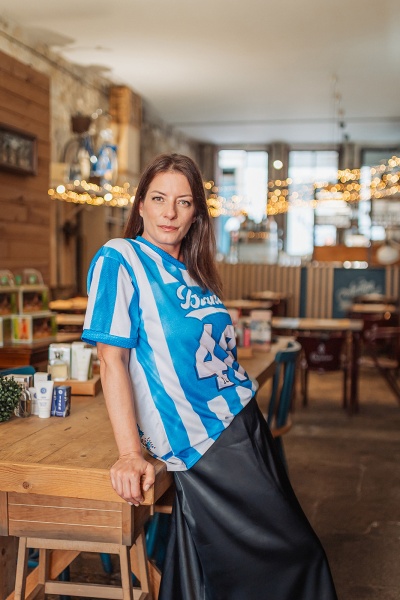 Mulher com t-shirt azul e branca e saia preta encostada a mesa de madeira num café