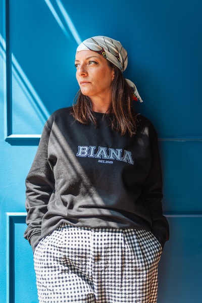 Mulher com lenço branco e camisola preta com texto BIANA EST. 2025 encostada a uma porta azul