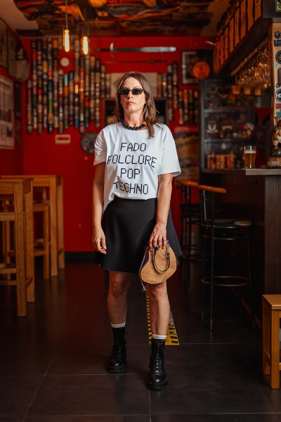 Mulher com camiseta branca com texto preto, saia preta, botas e bolsa pequena em cortiça num bar