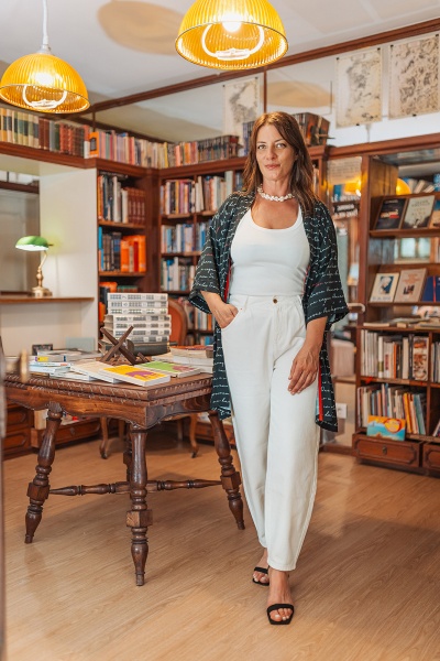 Mulher numa biblioteca com kimono preto, camisola branca e calças brancas ao pé de uma mesa de madeira com livros