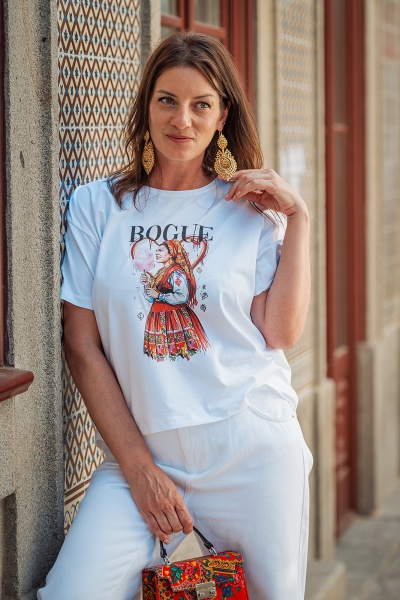 Mulher com t-shirt branca estampada, brincos dourados e mala vermelha floral junto a parede com azulejos