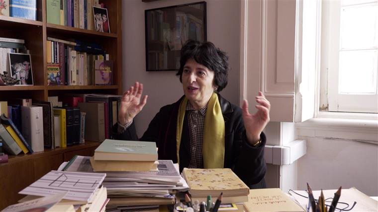 Pessoa sentada numa secretária com livros e cadernos empilhados, estante de livros ao fundo e janela iluminando a sala.